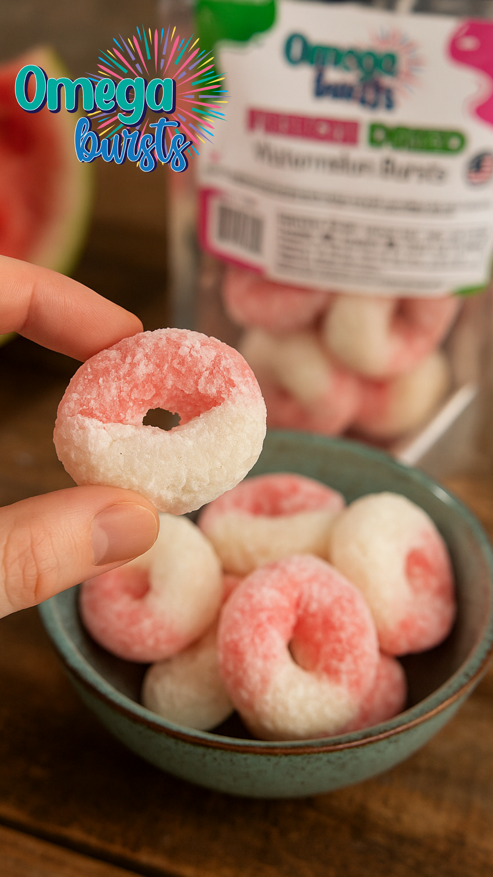Close-up of a hand holding a freeze-dried Omega Bursts Watermelon candy over a bowl with package in the background